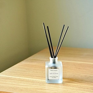 May include: A frosted glass essential oil diffuser with black reeds. The label reads "Salt & Citrus". The diffuser sits on a light wood surface. The background is a neutral color.