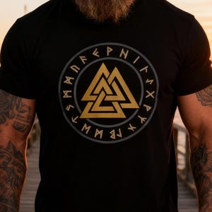 May include: Black t-shirt with a gold and grey circular design featuring a triangle symbol and runic text. The shirt has short sleeves and a crew neck. The design is centered on the chest.
