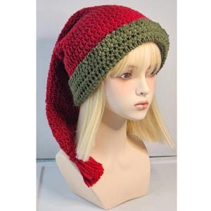 May include: A long, slouchy crochet hat featuring a vibrant red main body and a contrasting olive green folded brim. The hat has a pointed, elongated shape that hangs down to one side, finished with a small red tassel at the very tip.