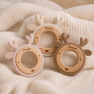 Könnte beinhalten: Drei kreisförmige Beißringe mit einem Holzkern und einem Silikonrand in Form eines Hirsches mit Geweih und Ohren. Jeder Ring ist mit einem Namen personalisiert: Paula in Hellrosa, Matteo in Creme und Samuel in Braun.