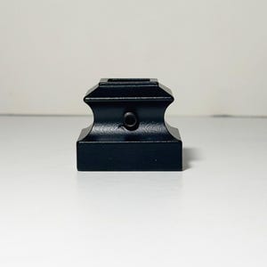 May include: A black, square-shaped decorative object with a stepped design. The object has a rectangular base and a square opening at the top. A small, circular hole is visible on the front. The object is made of a solid, matte black material.