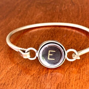 Pode incluir: Uma pulseira de metal em tom prateado com uma peça central circular que se assemelha a uma tecla de máquina de escrever vintage. O centro do componente circular preto exibe a letra E numa fonte de cor dourada, fixada por dois aros.