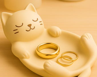May include: A cream-colored cat-shaped trinket dish holding three gold rings. The dish has a smiling cat face with closed eyes. The rings are of varying sizes. The dish is made of a smooth material.