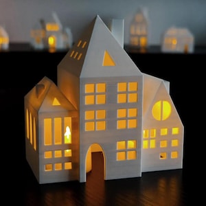 Könnte beinhalten: Ein weißer, mehrstöckiger Kerzenhalter in Form eines Hauses mit mehreren Fenstern und einem dreieckigen Dach. Die Fenster und Dachausschnitte leuchten mit warmem Licht, was darauf hindeutet, dass im Inneren Kerzen brennen. Weitere ähnliche Kerzenhalter befinden sich im Hintergrund.