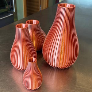 May include: Four copper-colored vases of varying sizes. Each vase has a ribbed, vertical design and a rounded, bulbous shape. The vases are arranged on a metallic surface, showcasing their unique textures and forms. The largest vase is approximately 10 inches tall.