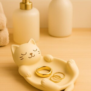 May include: A light yellow cat-shaped trinket dish holding three gold rings. The dish has a relaxed cat design with closed eyes and outstretched paws. A gold-colored soap dispenser and a white bottle are in the background.