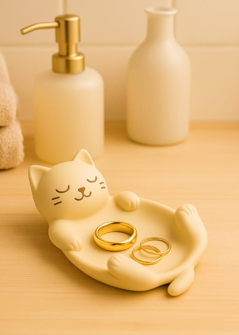 May include: A light yellow cat-shaped trinket dish holding three gold rings. The dish has a relaxed cat design with closed eyes and outstretched paws. A gold-colored soap dispenser and a white bottle are in the background.