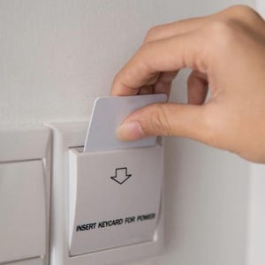 Könnte beinhalten: Eine weiße Keycard wird in einen weißen Netzschalter gesteckt. Der Schalter ist an einer weißen Wand montiert. Der Text "INSERT KEYCARD FOR POWER" ist auf dem Schalter aufgedruckt.
