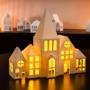 Könnte beinhalten: Ein weißer, papierförmiger Kerzenhalter in Form eines Hauses mit mehreren Fenstern und einem hohen Turm. Die Fenster leuchten mit warmem Licht, was auf Kerzen im Inneren hindeutet. Weitere ähnliche Häuser befinden sich im Hintergrund.