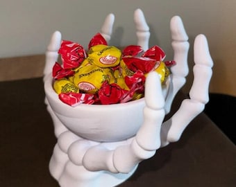 May include: A white, skeletal hand-shaped bowl filled with yellow and red wrapped candies. The bowl is a decorative item, perfect for Halloween or themed events. The candies are visible inside the bowl.