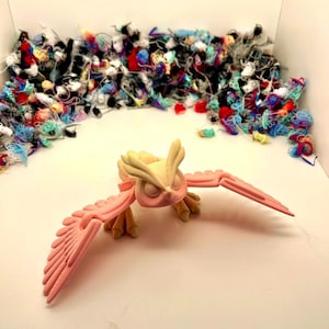 May include: A pink and cream-coloured 3D-printed owl figurine with its wings spread. The owl is set against a backdrop of colourful yarn scraps. The figurine features detailed feather and wing designs.