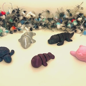 Könnte beinhalten: Eine Sammlung kleiner, farbenfroher Tierfiguren, darunter eine Schildkröte, ein Delfin, ein Alligator, eine Seekuh und ein Seehund. Die Figuren sind in Blau-, Silber-, Schwarz-, Lila- und Rosatönen gehalten. Im Hintergrund befindet sich ein Haufen bunter Wolle.