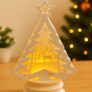 May include: A white, layered Christmas tree decoration with snowflake cutouts and a star on top. The tree features a warm, glowing interior scene with deer and trees. The base is a stacked, circular design.