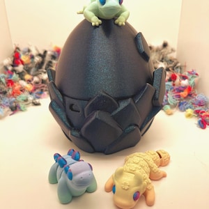 Könnte beinhalten: Eine dunkelblaue, eiförmige Skulptur mit strukturierter Oberfläche, gekrönt von einer hellgrünen Froschfigur. Zwei kleine, farbenfrohe Kreaturenfiguren befinden sich vor dem Ei. Eine ist blau und die andere gelb.