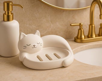 May include: A white ceramic soap dish shaped like a sleeping cat, with a matching soap dispenser with a gold pump. The bathroom accessories are on a beige countertop, with a gold faucet and mirror in the background.