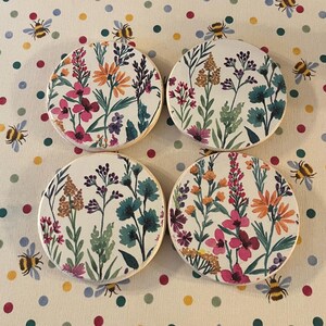May include: Four circular coasters with a floral design. Each coaster has a white background with colourful flowers in pink, orange, purple, and teal. The coasters are arranged on a dotted fabric.
