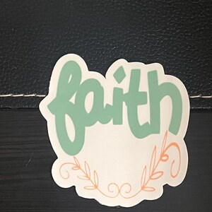 May include: A white sticker with the word "faith" written in a light green cursive font. The word is surrounded by a decorative border of orange leaves and swirls.