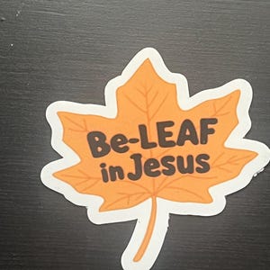 May include: A sticker in the shape of a fall leaf, colored orange with a white border. The sticker has the words "Be-LEAF in Jesus" in brown lettering.