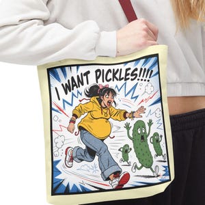 以下が含まれることがあります： クリーム色のトートバッグ。漫画風のイラストが描かれています。黄色いパーカーと青いジーンズを着た人が、漫画のピクルスに向かって走っている様子が描かれており、「I WANT PICKLES!!!!」という文字が入っています。バッグにはマルーン色のストラップが付いています。