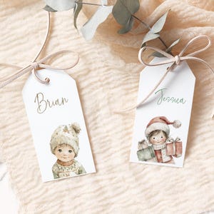 Pode incluir: Duas etiquetas de presente brancas com ilustra&ccedil;&otilde;es em aquarela de crian&ccedil;as em roupas de inverno. Uma etiqueta diz "Brian" e apresenta uma crian&ccedil;a com gorro e su&eacute;ter de malha. A outra diz "Jessica" e mostra uma crian&ccedil;a com gorro de Papai Noel segurando presentes.
