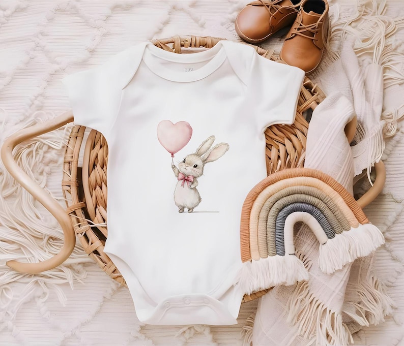 Puede incluir: Un body blanco para beb&eacute; con un globo en forma de coraz&oacute;n rosa y un conejito marr&oacute;n que sostiene el globo. El conejito lleva un lazo rosa.