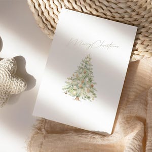 Pode incluir: Um cart&atilde;o de felicita&ccedil;&otilde;es branco com um desenho de &aacute;rvore de Natal em aquarela e a frase "Merry Christmas". O cart&atilde;o est&aacute; sobre uma superf&iacute;cie neutra com uma cesta tecida e um brinquedo em forma de estrela.
