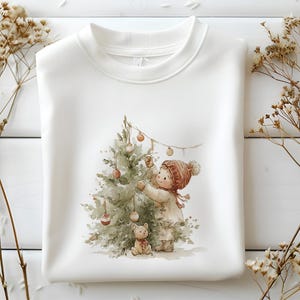 Pode incluir: Camisola branca com uma ilustra&ccedil;&atilde;o em aquarela de uma crian&ccedil;a decorando uma &aacute;rvore de Natal com enfeites. A crian&ccedil;a usa um chap&eacute;u e casaco vermelhos, com um ursinho de pel&uacute;cia por perto. Tema de f&eacute;rias aconchegante e festivo.