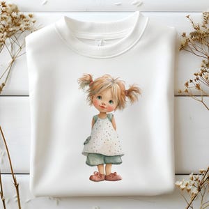 May include: White sweatshirt with a watercolour illustration of a young girl with blonde pigtails. She is wearing a white polka dot dress, light blue shorts, and pink shoes. The artwork has a soft, whimsical style.
