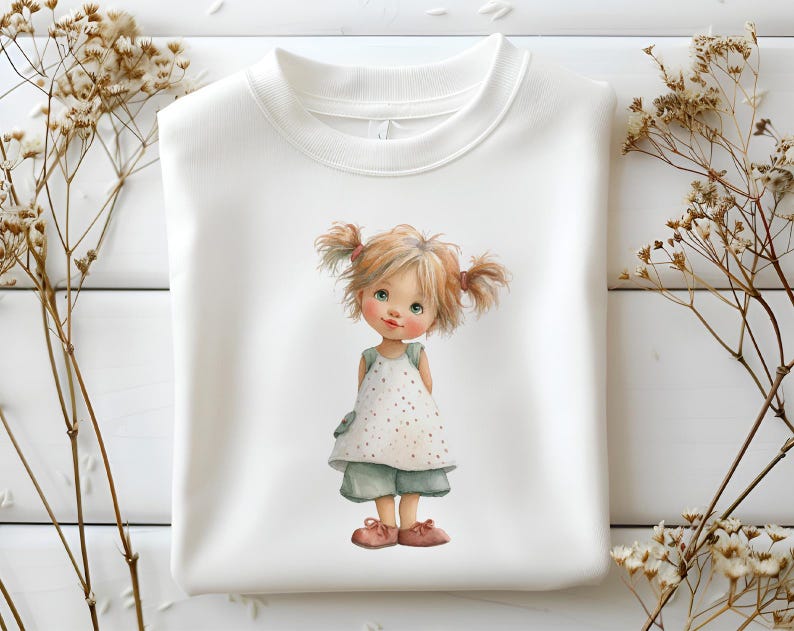 May include: White sweatshirt with a watercolour illustration of a young girl with blonde pigtails. She is wearing a white polka dot dress, light blue shorts, and pink shoes. The artwork has a soft, whimsical style.
