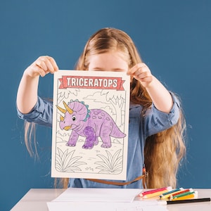 May include: A child holds a coloring page featuring a purple triceratops dinosaur with a yellow horn and blue accents. The word "TRICERATOPS" is printed at the top in a red banner. The background is a blue wall.