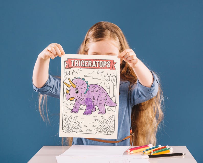 May include: A child holds a coloring page featuring a purple triceratops dinosaur with a yellow horn and blue accents. The word "TRICERATOPS" is printed at the top in a red banner. The background is a blue wall.