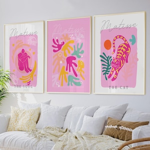 Op de afbeelding: Drie ingelijste kunstdrukken met gouden frames. Elke print heeft een roze achtergrond met abstracte ontwerpen en het woord "Matisse" in cursief. Eén print zegt "The Soul", een andere "The Cat".