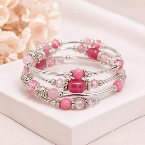 Può includere: Un bracciale avvolgente a più fili con filo metallico color argento e una varietà di perline rosa. Il design include perline rotonde lisce, perline di vetro sfaccettato e perline rosa cilindriche più grandi, con distanziatori argentati decorativi.