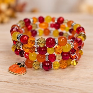 May include: A multi-strand wrap bracelet featuring a mix of red, orange, and yellow glass beads, faceted crystals, and silver-toned spacers. An orange pumpkin-shaped charm hangs from one end, and a small crystal dangle hangs from the other.
