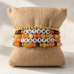 Puede incluir: Una pila de cuatro pulseras de cuentas con cuentas metálicas doradas, cuentas marrones facetadas y piedras lisas de color naranja, amarillo y crema. Dos pulseras incluyen cuentas de letras blancas que forman las palabras AUTUMN y THANKFUL.