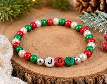 May include: A beaded bracelet with red, green, white, and silver beads. The word "JOY" is spelled out with letter beads. The bracelet is displayed on a wooden surface with Christmas decorations in the background.