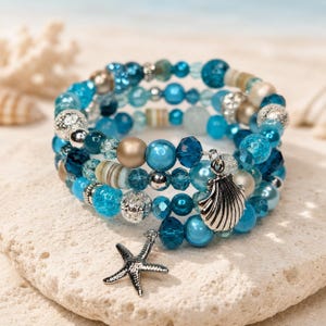 May include: A multi-strand wrap bracelet featuring a variety of blue, teal, and metallic gold beads in different textures and finishes. The bracelet includes a silver-toned metal seashell charm and a silver-toned metal starfish charm dangling.