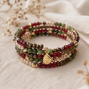 May include: A multi-strand wrap bracelet featuring alternating rows of deep red, olive green, metallic bronze, and cream-colored beads. The bracelet includes small gold-toned square spacers and a central gold-toned leaf-shaped charm pendant.