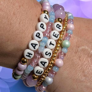 May include: Stack of beaded bracelets in pastel colors, including pink, blue, and lavender. One bracelet spells out "HAPPY EASTER" with white letter beads. Gold and iridescent beads add sparkle. A festive accessory.