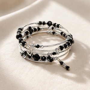 Könnte beinhalten: Ein Wickelarmband mit mehreren silberfarbenen Bändern. Das Armband ist mit schwarzen Perlen, kleinen silbernen Perlen und einem kleinen Herzanhänger verziert. Ein baumelnder Anhänger mit schwarzen Perlen hängt am Armband.