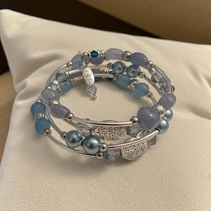 May include: A multi-strand bracelet with blue beads and silver accents. The bracelet features various shades of blue beads, including round, oval, and faceted shapes. Silver-toned metal wire and decorative charms complete the design. The bracelet is resting on a white surface.