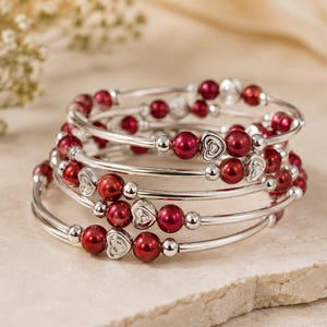May include: A stack of silver-toned, spiral bracelets. Each bracelet features red beads, silver beads, and heart-shaped charms. The bracelets are displayed on a light-colored, textured surface.