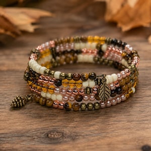 May include: A multi-strand wrap bracelet featuring a mix of cream, copper, and brown beads, including tiger eye stones and metallic accents. The bracelet is adorned with a small bronze-toned leaf charm and a miniature pinecone-shaped pendant.