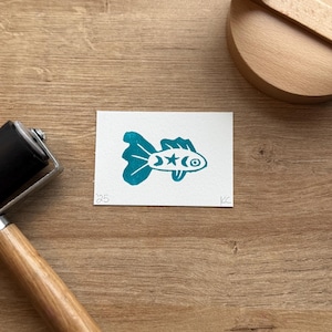 May include: A teal-colored fish print on white paper, featuring a star and crescent moon design. The print is on a wooden surface, with a wooden printing press and ink roller visible.