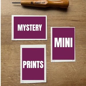 Puede incluir: Una herramienta Power Grip con mango de madera y punta metálica, encima de tres tarjetas rectangulares. Las tarjetas tienen un borde blanco y un centro morado oscuro con las palabras "MYSTERY", "MINI" y "PRINTS" en blanco.