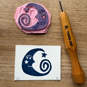 May include: A pink rubber stamp with a crescent moon and star design, a carved wooden handle tool, and a blue ink print of the same design on white paper. The stamp features a face within the moon and a spiral. The tool has "POWER GRIP" printed on it.