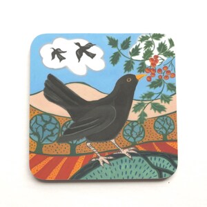 May include: A blackbird with a yellow beak perches on a green bush with red berries. Two blackbirds fly in the blue sky above a white cloud. The background is a colourful landscape with orange, brown, and green trees.