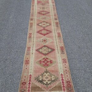 Puede incluir: Una alfombra larga y estrecha de estilo vintage con un patrón geométrico repetitivo en tonos de rosa, rojo, marrón y beige. La alfombra es rectangular y parece estar hecha de material tejido, adecuada para un pasillo o entrada.