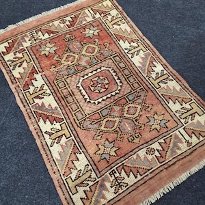 Puede incluir: Una alfombra rectangular tejida a mano con un detallado patrón geométrico en tonos rojos, beige y grises. La alfombra presenta un diseño de medallón central y un borde con flecos. La alfombra mide aproximadamente 91 cm de largo y 61 cm de ancho.