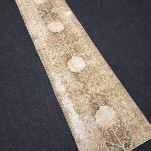May include: A rectangular, distressed vintage rug with a central floral design. The rug features a beige and brown color palette with accents of green. The design includes three large, circular floral motifs. The rug is placed on a dark surface.